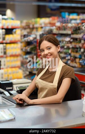 Tägliches Einkaufen. Junge Kassiererin im Supermarkt posiert mit der Kamera glücklich Stockfoto