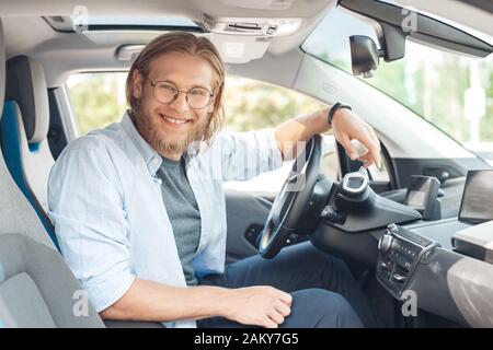Transport. Junger Mann in einer Brille, die mit dem Elektroauto unterwegs ist und in der Kamera fröhlich posiert Stockfoto