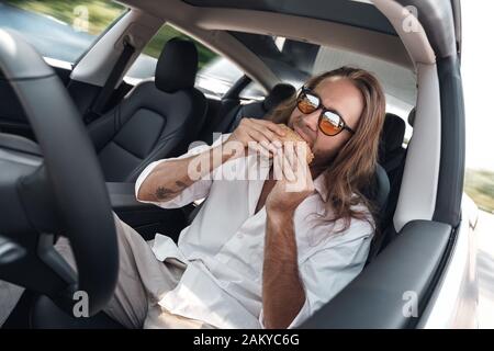 Transportmodus. Bärtiger Mann mit langen Haaren in der Sonnenbrille, der im Elektroauto sitzt und auf dem Autopiloten fährt, der Hamburger freudig isst Stockfoto