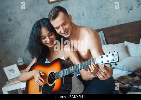 Gemischtes Rennpaar. Junger Mann, der Frau beigebracht hat, Gitarre auf dem Bett zu spielen, zusammen mit fröhlichem Gesang Stockfoto
