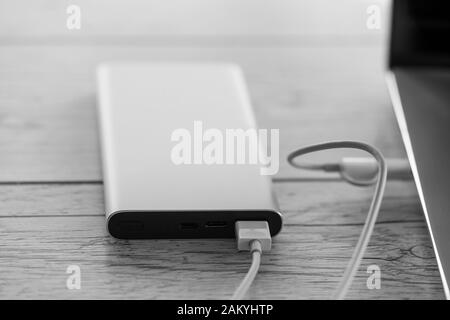 Aufladen des Laptops über eine Powerbank in silberfarbener Farbe, Ladebatterie auf Holzplatten im Hintergrund. Produktives tragbares Gerät für Reisen, Schwarzweiß Stockfoto