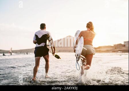 Fröhliche Surfer, die bei Sonnenuntergang im Wasser laufen - Junge Paare haben Spaß beim Surfen im Meer - Lifestyle-Konzept für Extremsport und Jugendkultur Stockfoto