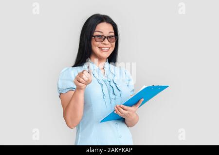 Freistil. Asiatische Frau in Brillen, isoliert auf Weiß stehend, mit Zwischenablage, die auf Kamera verspielt zeigt Stockfoto