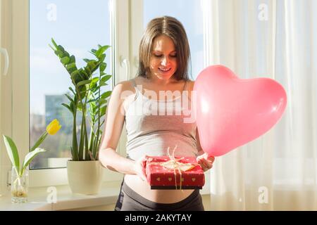 Urlaub im Leben der jungen, schönen schwangeren Frau, glückliche Frau mit Valentinsgeschenkkasten und rotem Herzballon. Mutterschaft und Schwangerschaft Stockfoto