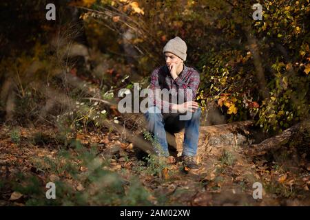 Trauriger Teenager in Mütze, Flanell und Jeans sitzt auf Holzholz. Stockfoto