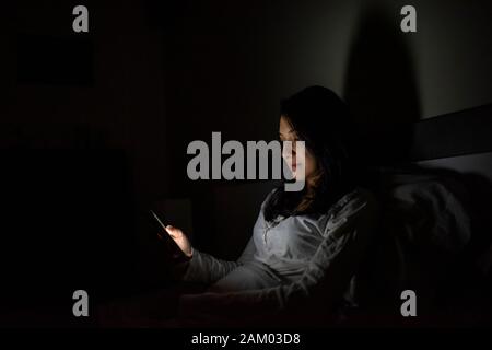 Dunkelhaarige Latina Frau im Schlafanzug können Sie über Ihr Handy im Bett liegend Stockfoto