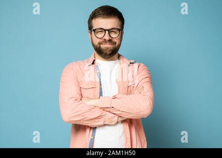 Lustiger bärtiger Mann mit Brille steht mit gekreuzten Armen, lächelt seltsam und blickt mit verengten Augen auf die Kamera. Stockfoto