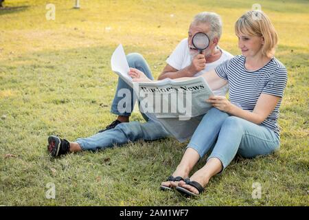 Der ältere Mann, der eine Lupe hält, um eine Zeitung mit seiner Frau neben ihm zu sehen, das ältere Paar sitzt, um Zeitungen auf dem Rasen in der PA zu sehen Stockfoto