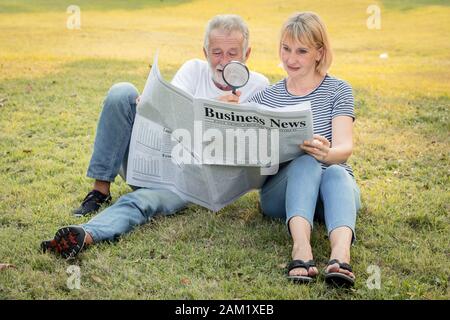 Der ältere Mann, der eine Lupe hält, um eine Zeitung mit seiner Frau neben ihm zu sehen, das ältere Paar sitzt, um Zeitungen auf dem Rasen in der PA zu sehen Stockfoto