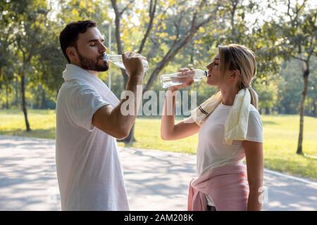 Junge Paare, die nach dem Training im Park am Morgen Wasser trinken. Konzept Paare trainieren im Park. Frauen und Männer heben Wasserflaschen an und Stockfoto