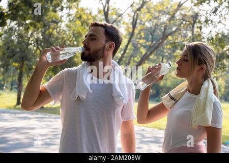 Junge Paare, die nach dem Training im Park am Morgen Wasser trinken. Konzept Paare trainieren im Park. Frauen und Männer heben Wasserflaschen an und Stockfoto