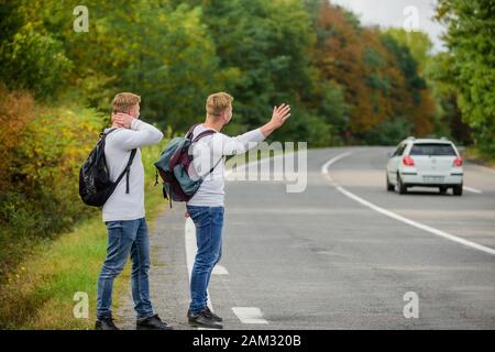 Neue Leute zu treffen. Auf der Suche nach transportieren. Zwillinge zu Fuß entlang der Straße. stop Auto mit "Daumen hoch" Geste. per Anhalter fahren und anhalten Auto mit Daumen nach oben Geste auf die Landschaft. Auf der Straße. Genießen Sommer wandern. Stockfoto