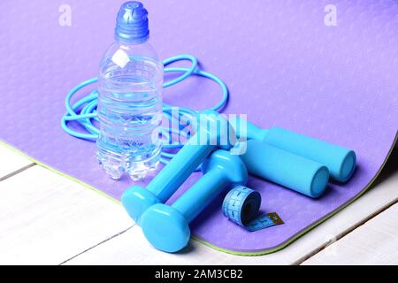 Shaping und Fitness. Barbells und Springseil neben der Wasserflasche auf der lila Yogamatte. Sport und gesundes Lifestyle-Konzept. Hanteln, Maßband und Springseil in zyanfarbener Farbe auf Holzhintergrund Stockfoto