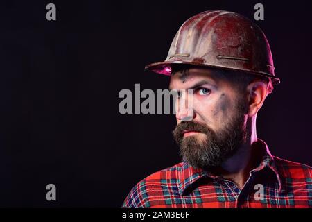 Mann mit misstrauischem Gesicht auf schwarzem Hintergrund, Kopierbereich. Erbauer oder Bergmann mit dickem Bart. Arbeiter mit brutalem Bild trägt schmutzigen Helm und Krawthemd. Bau- und Arbeitskonzept Stockfoto