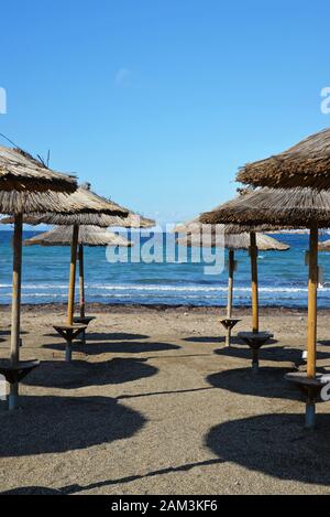 Stroh Sonnenschirme in Zeilen werfen Schatten auf dem Sand an einen leeren Strand im Winter, bei Vouliagmeni, Athen, Griechenland. Stockfoto