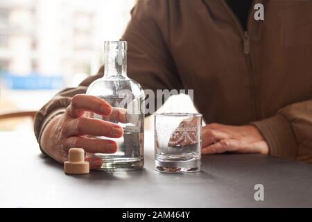 Mann, der eine Flasche Alkohol und ein leeres Glas nebeneinander an einem Tisch in der Heim- oder Barumgebung hält. Selektive Fokussierung auf Hand und Flasche. Clos Stockfoto