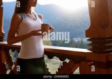 Die schlanke kaukasische Frau hält eine Tasse Tee in ihren Händen im Bergresort. Sportmädchen mit heißem Kaffeebecher auf dem Holzbalkon des Landhauses. Wald und Stockfoto