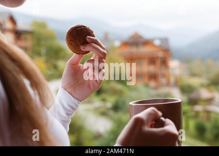 Frau hält eine Tasse Kaffee und Haferplätzchen in ihren Händen im Bergresort. Frau mit Teebecher und Snack mit Wald und Holzhotel im Hintergrund Stockfoto