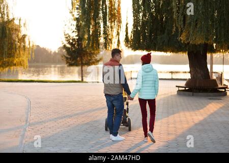 Rückansicht des jungen, liebevollen Paares, das die Hände hält. Eltern mit Kinderwagen im Herbstpark. Liebe, Elternschaft, Familie, Saison und Menschen Konzept. Stockfoto