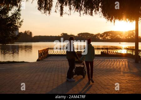 Rückansicht junger, liebevoller Paar-Silhouetten, die die Hände halten. Eltern mit Kinderwagen im Herbstpark bei Sonnenuntergang. Liebe, Elternschaft, Familie, Saison und Stockfoto