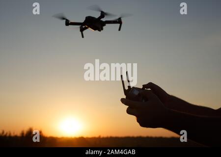 Der Mann hält die Fernbedienung mit den Händen, während der Kopter vor dem Hintergrund fliegt. Drohne schwebt hinter dem Piloten auf Suset. Kein Gesicht Stockfoto