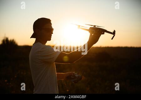 Silhouette eines Mannes, der kleine kompakte Drohne und Fernbedienung in den Händen hält. Pilot startet Quadcopter aus seiner Handfläche bei Sonnenuntergang. Drohne bereit zu Stockfoto