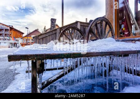 Holztisch mit Eis und hängenden Eiszapfen bedeckt. Holzkarren, Radschmuck in Les deux Alpen, Alpendorf in Frankreich Stockfoto