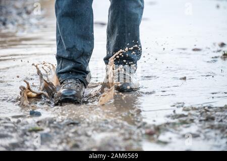 Mann in robusten braunen Wanderstiefeln, die nach starkem Regen im Winter auf einem Weg durch eine tiefe schlammige Schlammpudel spritzen Stockfoto