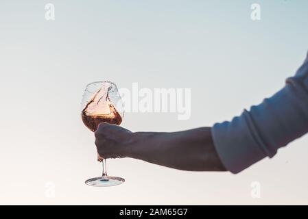 Glas Rosengwein mit Spritzern in der Hand der Frau gegen den Himmel bei Sonnenuntergang. Stockfoto