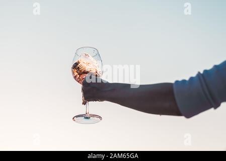 Glas Rosengwein mit Spritzern in der Hand der Frau gegen den Himmel bei Sonnenuntergang. Stockfoto