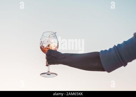 Glas Rosengwein mit Spritzern in der Hand der Frau gegen den Himmel bei Sonnenuntergang. Stockfoto