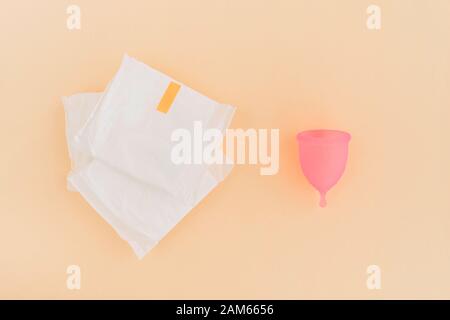 Weibliche Hygieneprodukte - Menstruationsbecher und Stapel von Sanitärpolstern auf hellorangefarbenem Hintergrund. Stockfoto