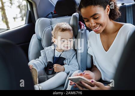 Junge Mutter mit ihrer Tochter sitzt auf einem Rücksitz eines Autos. Frau und ein kleines Mädchen, die auf dem Handy im Auto suchen. Stockfoto