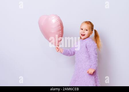 Glückliches Herz des Valentinstag. Kleines Mädchen lächelt mit rosafarbenem Ballonherz auf hellviolettem Hintergrund. Stockfoto