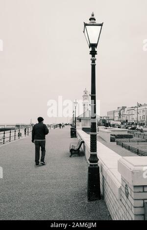 Herne Bay, Großbritannien - 8. Januar 2020. Monochromes Bild einer Straßenleuchte im Vintage-Stil an der Herne-Bay-Promenade, als ein Mann zum Uhrturm vorbeizieht. Stockfoto