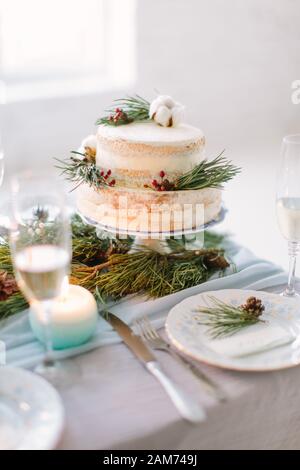 Hochzeitstorte mit Pinien, Beeren und Baumwollblumen auf dem Hochzeitstisch mit Dekor, Platten und Kerzen Stockfoto
