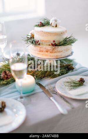 Hochzeitstorte mit Pinien, Beeren und Baumwollblumen auf dem Hochzeitstisch mit Dekor, Platten und Kerzen Stockfoto