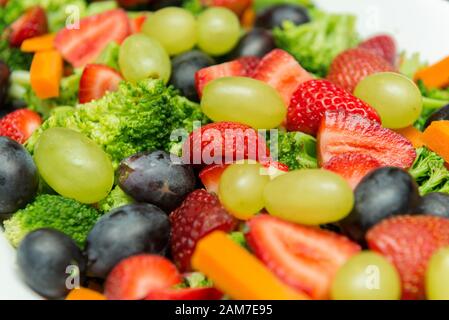 Nahaufnahme von Obstsalat mit Brokkoli, grünen und schwarzen Trauben, Erdbeeren- und Karottenscheiben oder Würfeln. Köstliche, nahrhafte und gesunde Mahlzeit in einem Whit Stockfoto