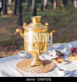 Goldener russischer Samowar mit Teetassen und süßen Snacks auf dem Tisch im Freien Stockfoto