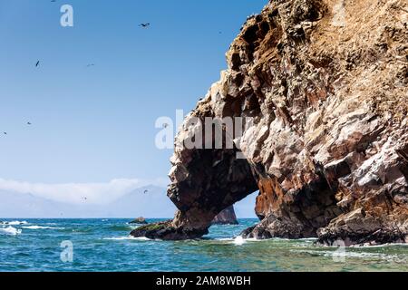 Ein Felsen mit einer Grotte mit Blick auf das Meer, Möwen fliegen herum, Berge, Ballestas-Inseln sind in der Ferne zu sehen, Paracas Rum, Peru, Latin Stockfoto