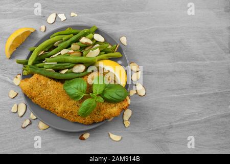 Parmesan verkrustete Tilapia mit einem Sprig frischen Basilikums. Grüne Bohnen, in Scheiben geschnittene Mandeln und Zitronen zieren den Teller in dieser mediterranen Mahlzeit. Stockfoto