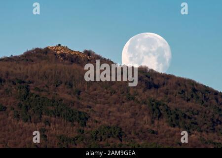 Der Vollmond setzt früh am Morgen hinter einem Berg gegen den blauen Himmel Stockfoto