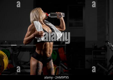 Seitenansicht der blonden Frau im schwarzen Sportoutfit im Fitnessstudio. Ernte einer muskulösen, tätowierten Frau mit weißem Handtuch auf dem schulterigen Trinkwasser und nach dem Training Ruhe. Konzept Bodybuilding. Stockfoto