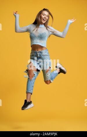 Vorderansicht des fröhlichen Mädchens in voller Höhe mit langen braunen Haaren, die das linke Bein im Sprung nach oben heben und ein kurzes blaues Oberteil mit langen Ärmeln und gerippten Jeans tragen. Stockfoto