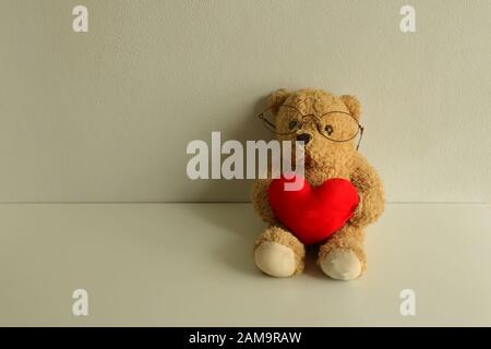 Teddybär-Puppe mit Brillen auf weißem Holztisch, der rotes Herz hält und Kamera, Liebeskonzept betrachtet Stockfoto