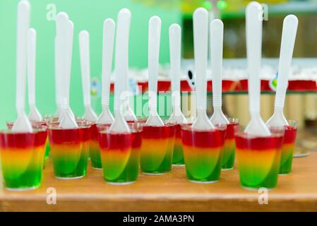 Glas mit farbenfroher gestreifter Gelee über Holztisch. Rainbow Jelly für Kinder Party. Löffel aus Kunststoff in Gelee. Köstliches Dessert Für Die Kinderparty. Platz Stockfoto