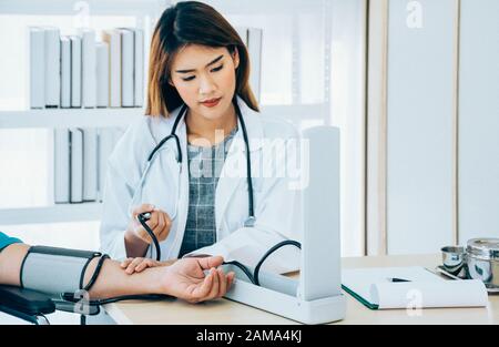 Junge schöne asiatische Ärztin misst den Blutdruck für einen Patienten, der auf einem Rollstuhl sitzt, indem sie ein Blutdruckmessgerät in einem Krankenhaus verwendet Stockfoto