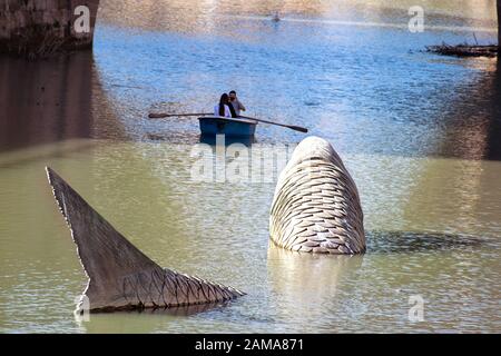 Murcia, Murcia, Spanien. Januar 2020. Riesige Fischskulptur im Fluss Segura gegen Menschen, die auf dem Boot rasten. Stockfoto