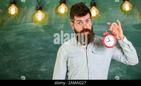 Lehrer in Brillen hält Wecker. Schulzeitplan Konzept. Mann mit Bart und Schnurrbart auf überraschtem Gesichtsstand im Klassenzimmer. Bärtiger Hüpfer hält Uhr, Schwarzen Brett im Hintergrund, Kopierraum. Stockfoto