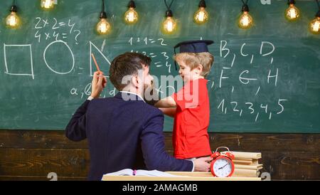 Der junge männliche Lehrer erklärt das Thema, führt seinen Schüler zum Lernen, während er im Klassenzimmer sitzt, Kelkbrett mit Scribbles auf dem Hintergrund, Rückansicht. Stockfoto